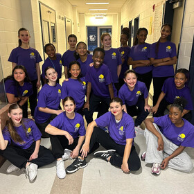 Groupe d’élèves souriantes posant dans un corridor d’école, vêtues de t-shirts violets de l’Académie des arts, certaines debout et d’autres accroupies, après une activité artistique.